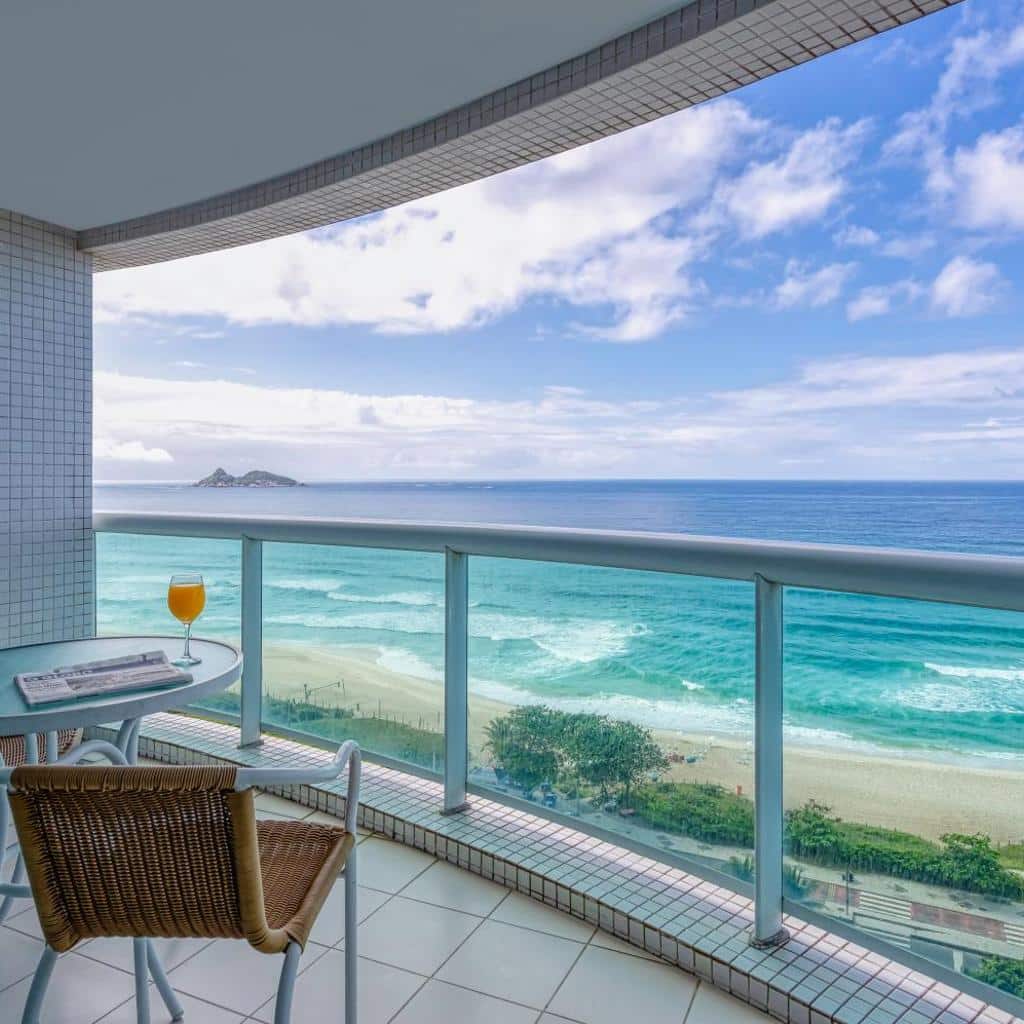 Uma varanda no Wyndham Rio Barra com cadeira, mesa e suco de laranja tem vista para a praia de areia e o oceano azul.
