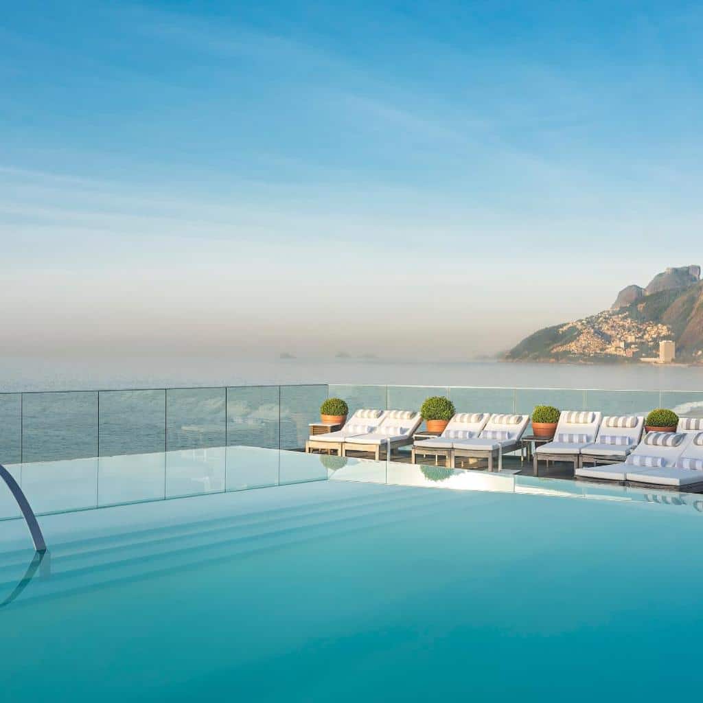 Piscina infinita no Hotel Fasano Rio de Janeiro com espreguiçadeiras, vista para o mar e montanhas distantes sob um céu azul claro.
