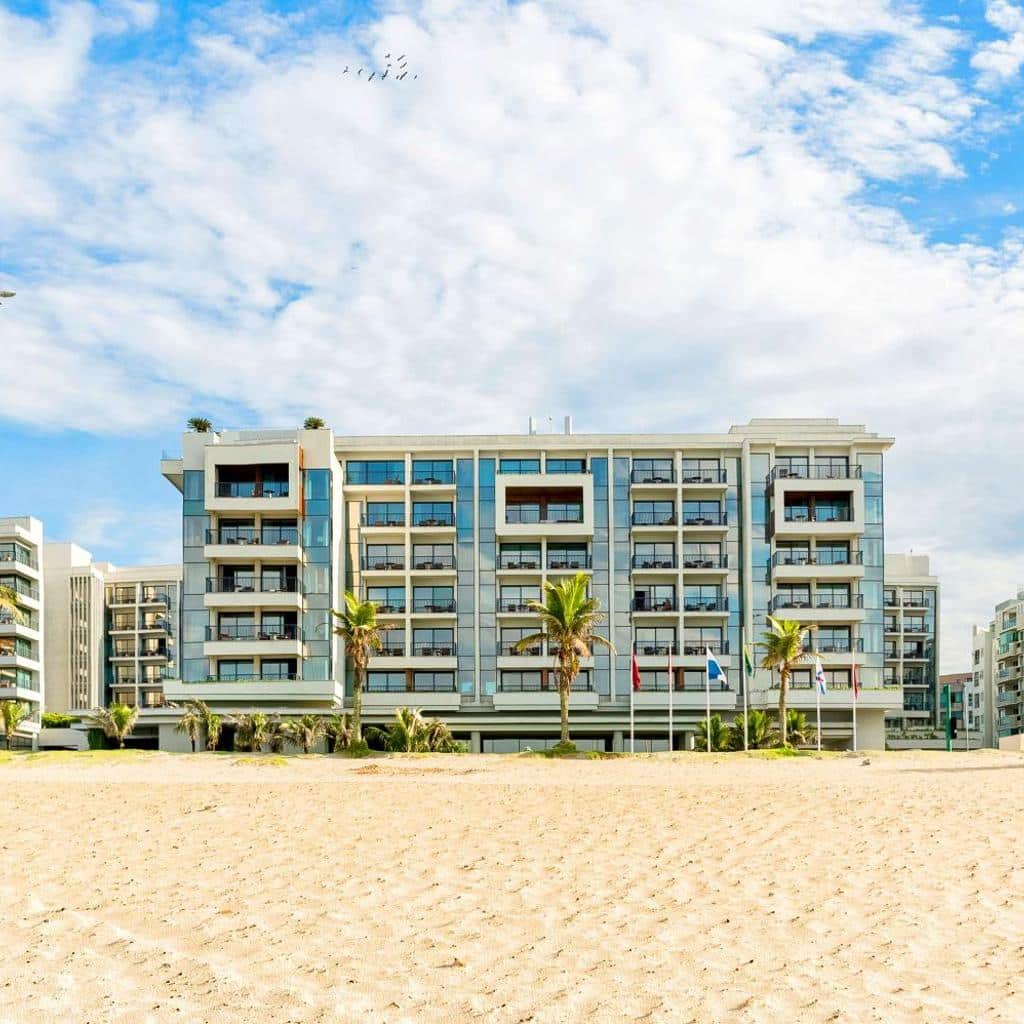 Moderno hotel Grand Hyatt Rio de Janeiro à beira-mar com varandas e palmeiras, visto de uma praia de areia sob um céu nublado.