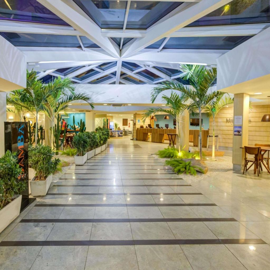Lobby do ibis Rio de Janeiro Barra da Tijuca com grandes vasos de plantas, teto com claraboia, piso de azulejos e balcão de recepção.
