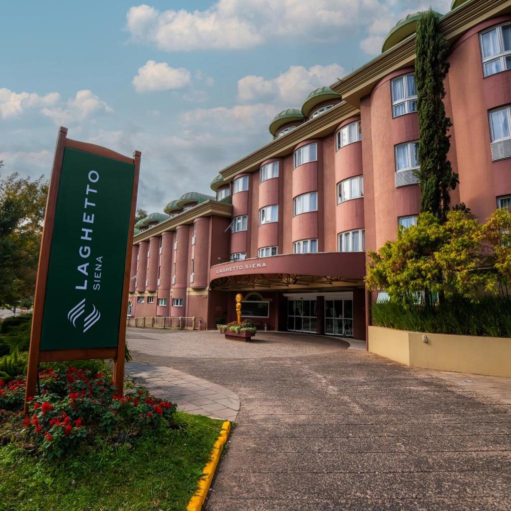 Hotel Laghetto Siena Gramado, curvo e de vários andares, com letreiro, jardim e céu nublado ao fundo.