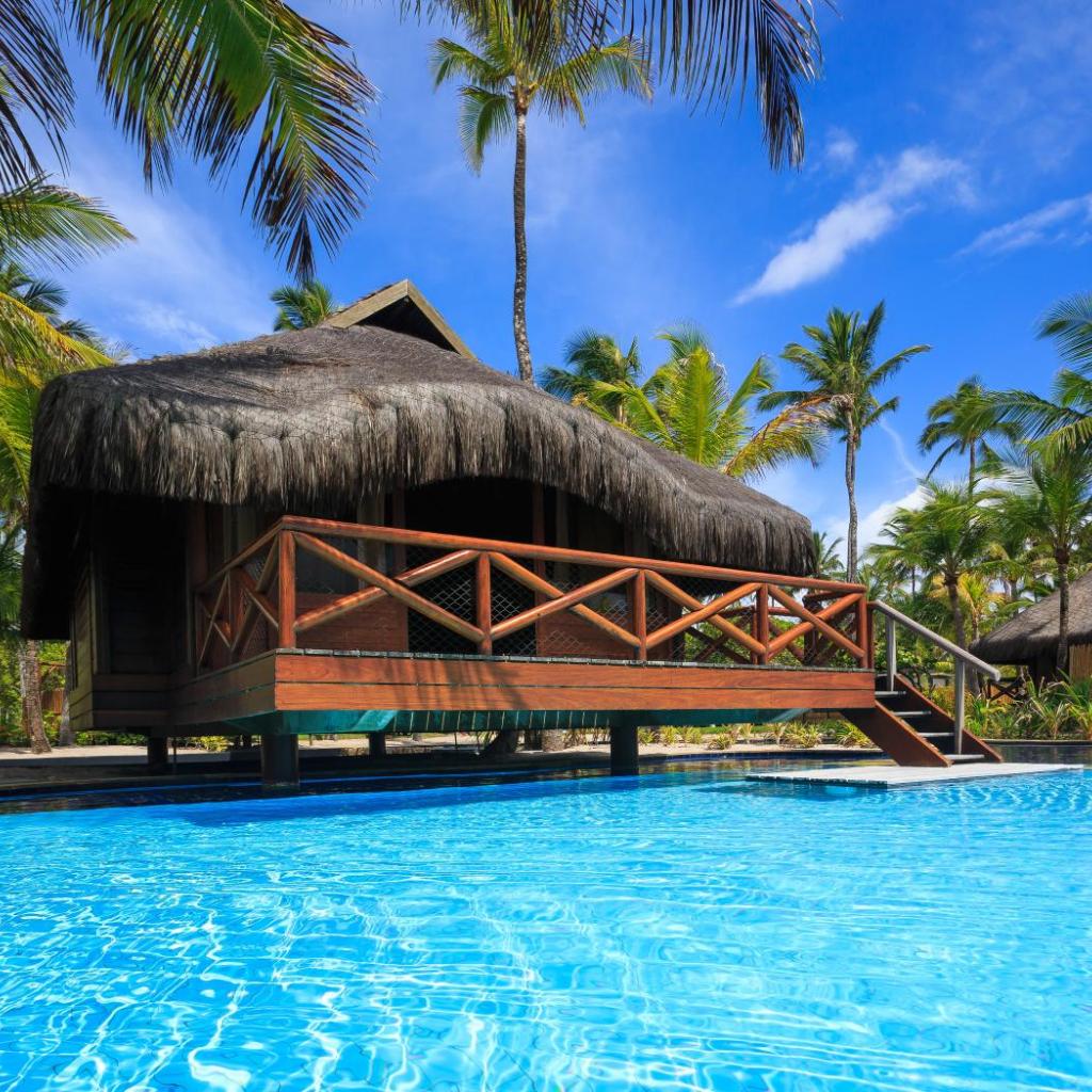 Bangalô tropical com telhado de palha ao lado de uma piscina azul clara em NANNAI Muro Alto, cercado por palmeiras.