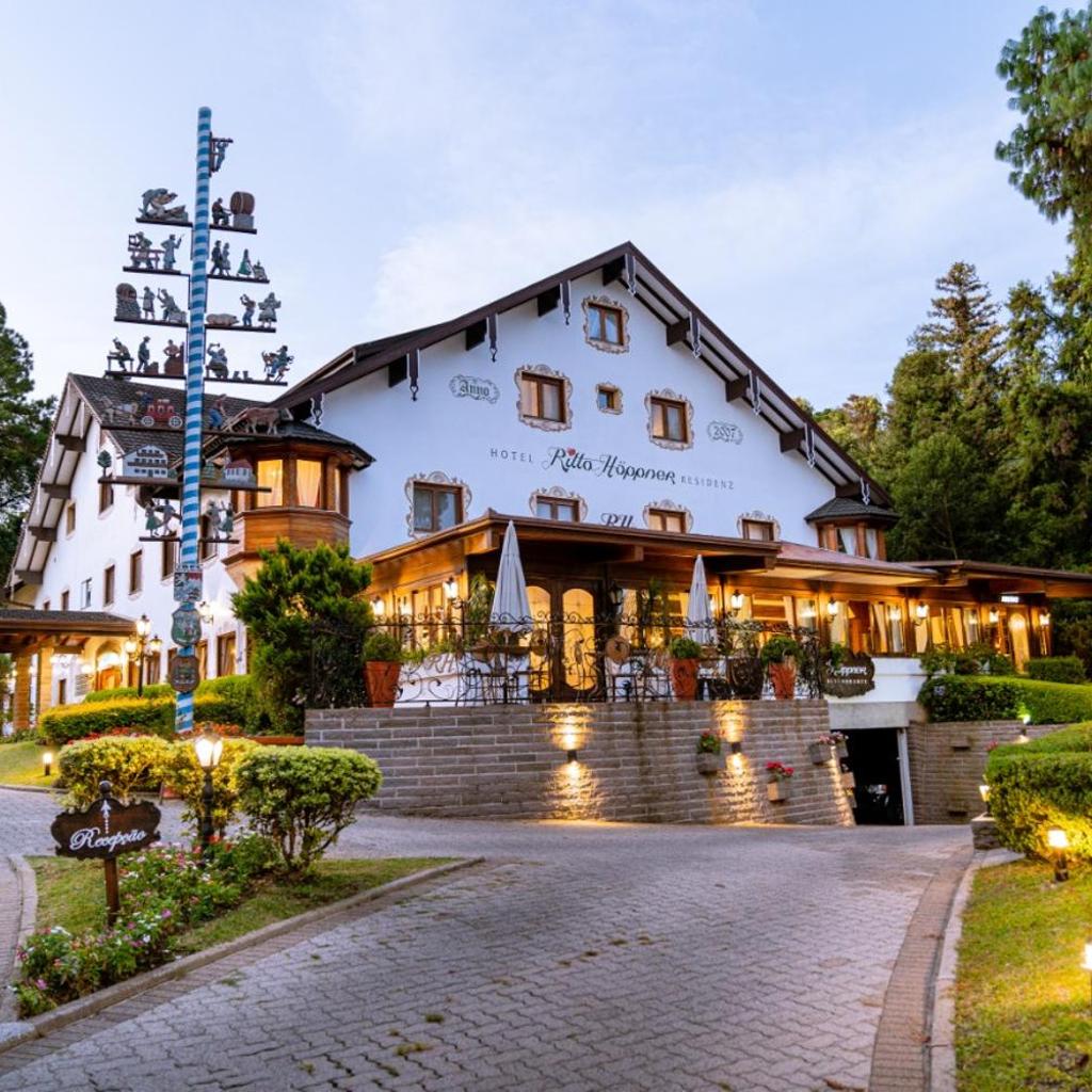 Hotel Ritta Höppner, em estilo alpino, com paredes brancas, acabamento marrom, guarda-chuvas no pátio e paisagismo no jardim ao anoitecer.