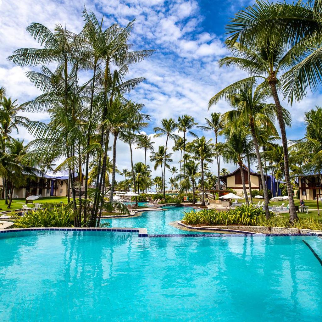 Uma piscina tropical do Summerville Resort - All Inclusive cercada por palmeiras, espreguiçadeiras e um céu azul com nuvens.