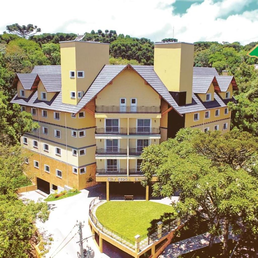Sky Serra Hotel: Um grande hotel amarelo com um telhado cinza, cercado por árvores e vegetação sob um céu parcialmente nublado.